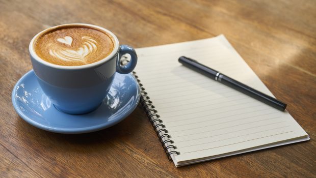 A cup of coffee with a blank notebook and pen next to it.