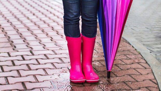 A pair of women's legs in rain boots with a pink and purple umbrella next to them.