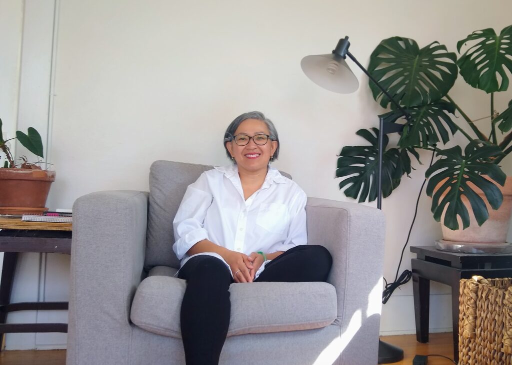 Middle-age Asian woman, wearing white shirt and black pants, sitting on swivel couch in living room and surrounded by house plants.