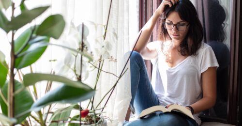 A young woman reading a book while sitting on a ledge near the window.