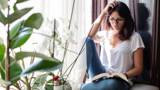 A young woman reading a book while sitting on a ledge near the window.